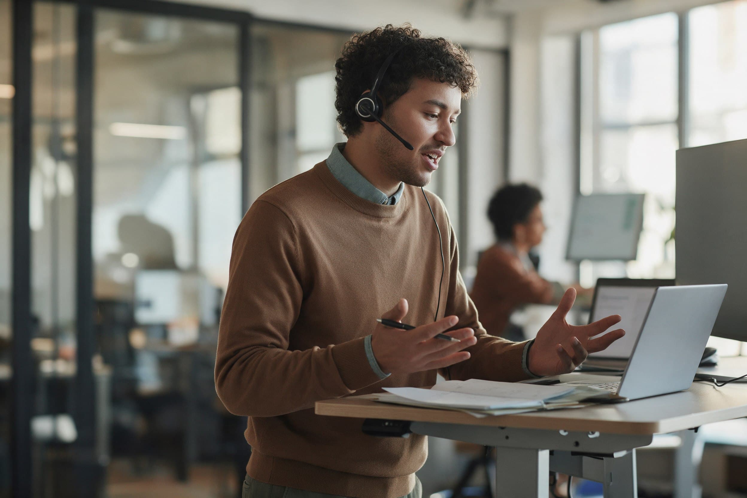 An account manager on a call at a standing desk with a pipeline board behind, mid conversation
